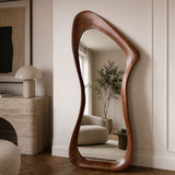 An organic-shaped full-length floor mirror with a rich, wavy brown wooden frame, styled in a warm, modern living room setting.