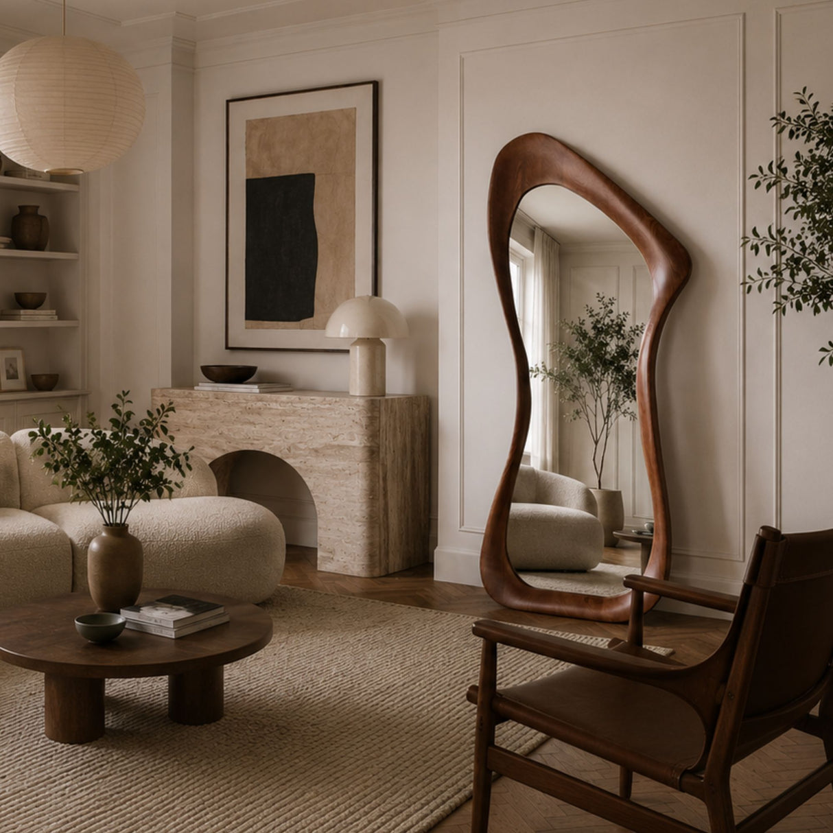 A modern living room scene featuring a tall, wavy wooden floor mirror with an organic, fluid silhouette. The mirror is styled in a warm, minimalist interior with a beige sofa, a round wooden coffee table, and a stone fireplace in the background.