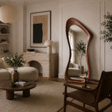 A modern living room scene featuring a tall, wavy wooden floor mirror with an organic, fluid silhouette. The mirror is styled in a warm, minimalist interior with a beige sofa, a round wooden coffee table, and a stone fireplace in the background.