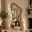 An organic-shaped full-length floor mirror with a natural oak wood frame stands in a sunlit room next to a fireplace and large potted plants.