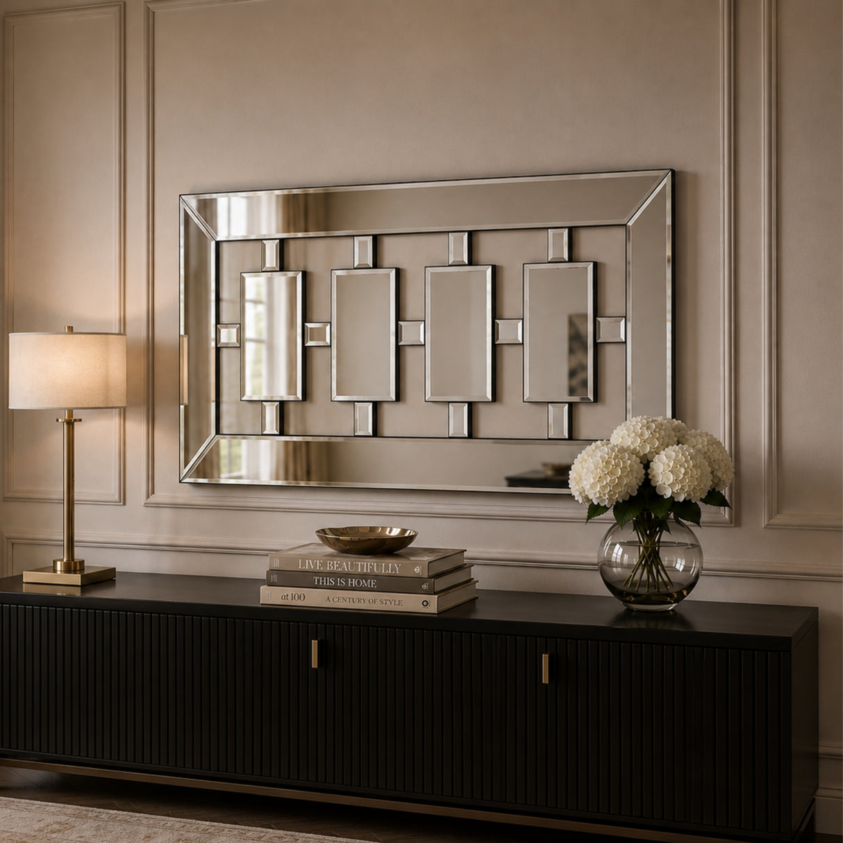 A rectangular wall mirror with a geometric frame and mirrored panels, mounted on a beige paneled wall above a black console table with a lamp, books, and a vase of white flowers.