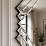 A modern geometric wall mirror with a black zigzag frame and reflective silver mirrored panels, mounted on a white paneled wall in a styled interior room.
