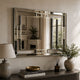 A large rectangular wall mirror with a polished silver frame hangs on a beige wall above a dark wooden console table. The scene includes a table lamp, books, a small diffuser, and potted plants, with the mirror reflecting an interior fireplace and window view.