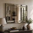 A large rectangular wall mirror with a polished silver frame hangs on a beige wall above a dark wooden console table. The scene includes a table lamp, books, a small diffuser, and potted plants, with the mirror reflecting an interior fireplace and window view.