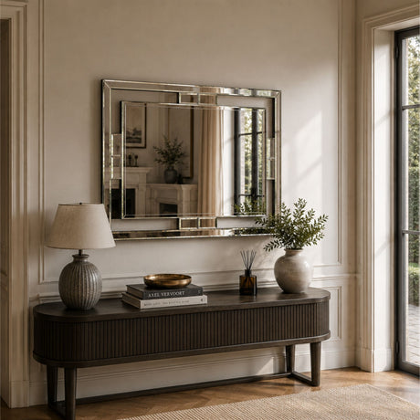 A large rectangular wall mirror with a polished silver frame hangs above a dark wood console table in a bright, elegant room with beige walls, a table lamp, books, and a vase.