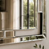A close-up of a rectangular wall mirror with a polished silver-toned frame, reflecting a softly blurred interior and window view.