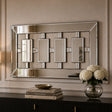 A rectangular wall mirror with a geometric silver-toned frame and four central rectangular mirror panels, mounted on a beige wall above a dark console table with books and a vase of white flowers.