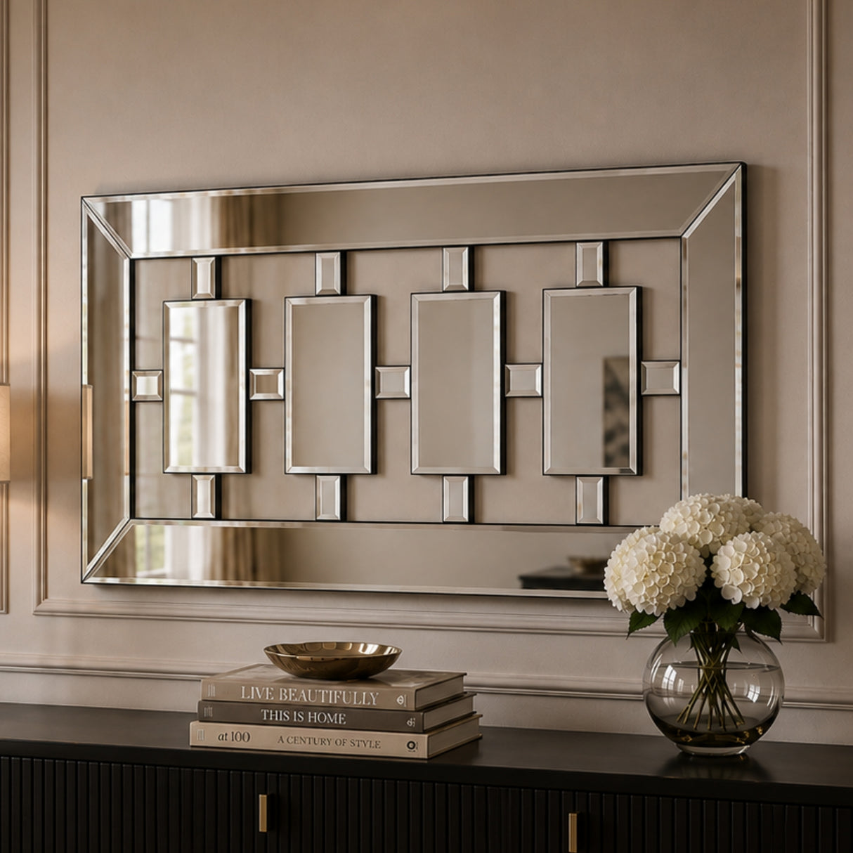 A rectangular wall mirror with a geometric silver-toned frame and four central rectangular mirror panels, mounted on a beige wall above a dark console table with books and a vase of white flowers.