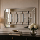 A rectangular wall mirror with a geometric silver-toned frame and four central rectangular mirror panels, mounted on a beige wall above a dark console table with books and a vase of white flowers.