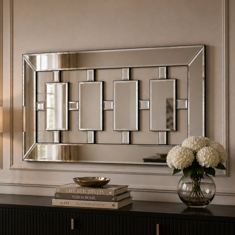 A rectangular wall mirror with a geometric silver-toned frame and four central rectangular mirror panels, mounted on a beige wall above a dark console table with books and a vase of white flowers.