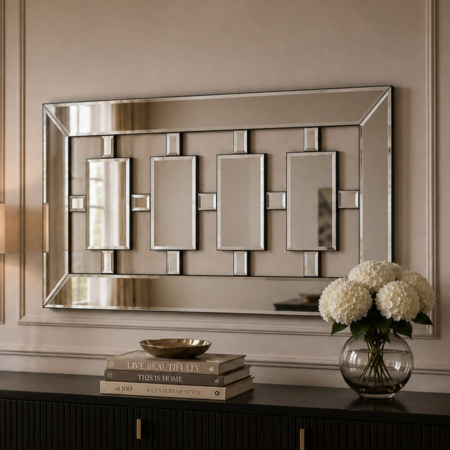 A rectangular wall mirror with a geometric silver-toned frame and four central rectangular mirror panels, mounted on a beige wall above a dark console table with books and a vase of white flowers.