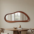 Wooden mirror with wavy design on a beige wall above a wooden table with decor items.