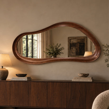 An organic-shaped wall mirror with a warm brown wooden frame hanging above a wooden console table in a styled room setting.