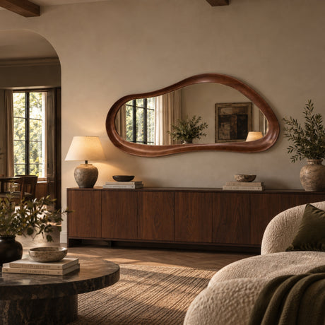An organic-shaped wooden-framed mirror hangs on a beige wall above a dark wood sideboard in a styled living room. The scene includes a lamp, books, plants, a sofa, and a round coffee table, with warm natural light coming from a window on the left.