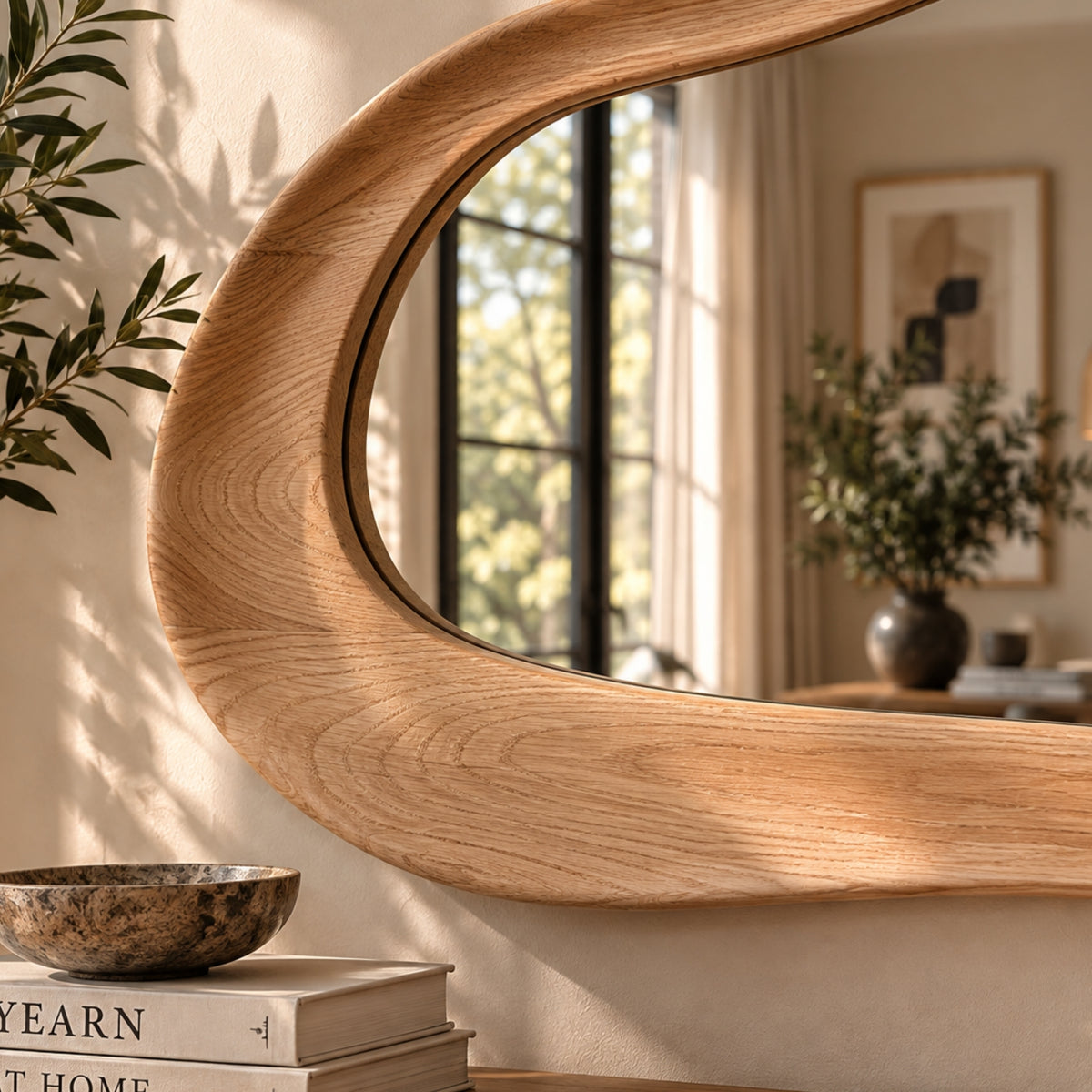 A large, organic-shaped wall mirror with a thick, curved light oak wood frame, mounted on a beige wall in a sunlit room. The reflection shows a window and curtains, and the surrounding environment includes a decorative bowl on books and a potted plant.