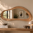 An organic-shaped wooden wall mirror with a thick, flowing frame, mounted on a beige wall above a wooden console table decorated with books, bowls, and a lamp.