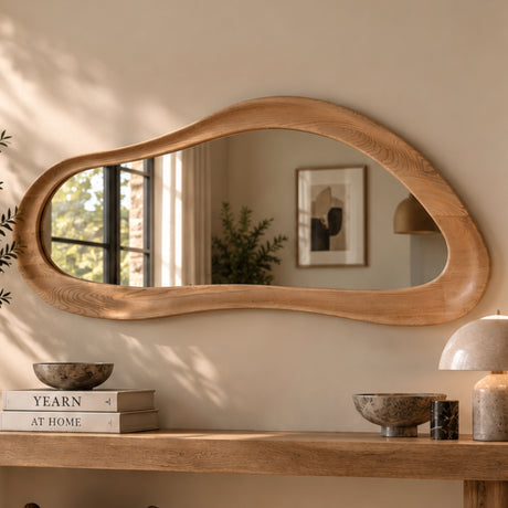 An organic-shaped wooden wall mirror with a thick, flowing frame, mounted on a beige wall above a wooden console table decorated with books, bowls, and a lamp.