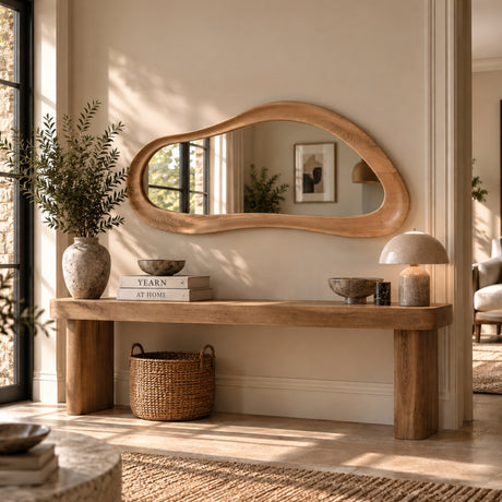 An organic-shaped wooden wall mirror with a natural oak frame hangs above a rustic wooden console table in a sunlit, modern entryway. The room features neutral beige walls, a woven basket, a ceramic vase with greenery, and a mushroom-shaped table lamp.