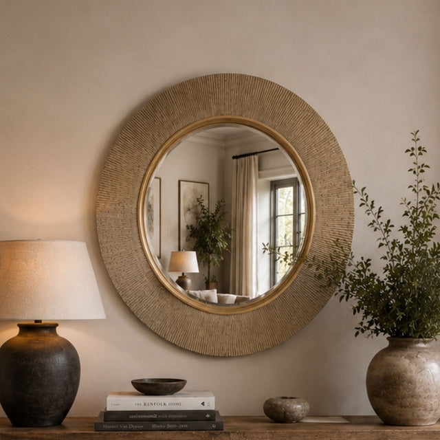 Round wall mirror with a textured beige woven frame and a gold-toned inner border, displayed on a console table in a styled room setting with a lamp, books, and a potted plant.