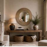 A round woven wood mirror hangs on a beige wall above a rustic wooden console table in a styled living room. The table holds a dark ceramic table lamp, stacked books, and a small decorative object, with a large textured vase of greenery on the right.