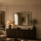A large arched wall mirror with a distressed gold frame hangs above a dark wood console in a warm, beige-toned living room. The mirror reflects a window and greenery, while the room also features a table lamp, books, and a vase with dried branches.