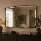 A large arched wall mirror with a thin gold frame hangs above a dark wood console table in a warm, neutral-toned room. The mirror reflects a window, curtains, and framed art, while the room itself is styled with a lamp, books, and a vase with greenery.