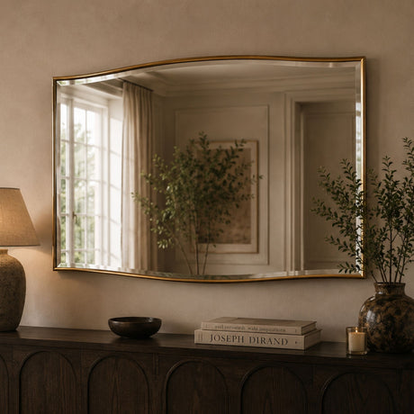 A large arched wall mirror with a thin gold frame hangs above a dark wood console table in a warm, neutral-toned room. The mirror reflects a window, curtains, and framed art, while the room itself is styled with a lamp, books, and a vase with greenery.