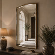 A large arched wall mirror with a gold frame hangs on a beige wall above a console table. The mirror reflects a softly lit hallway with an arched window and potted plants.