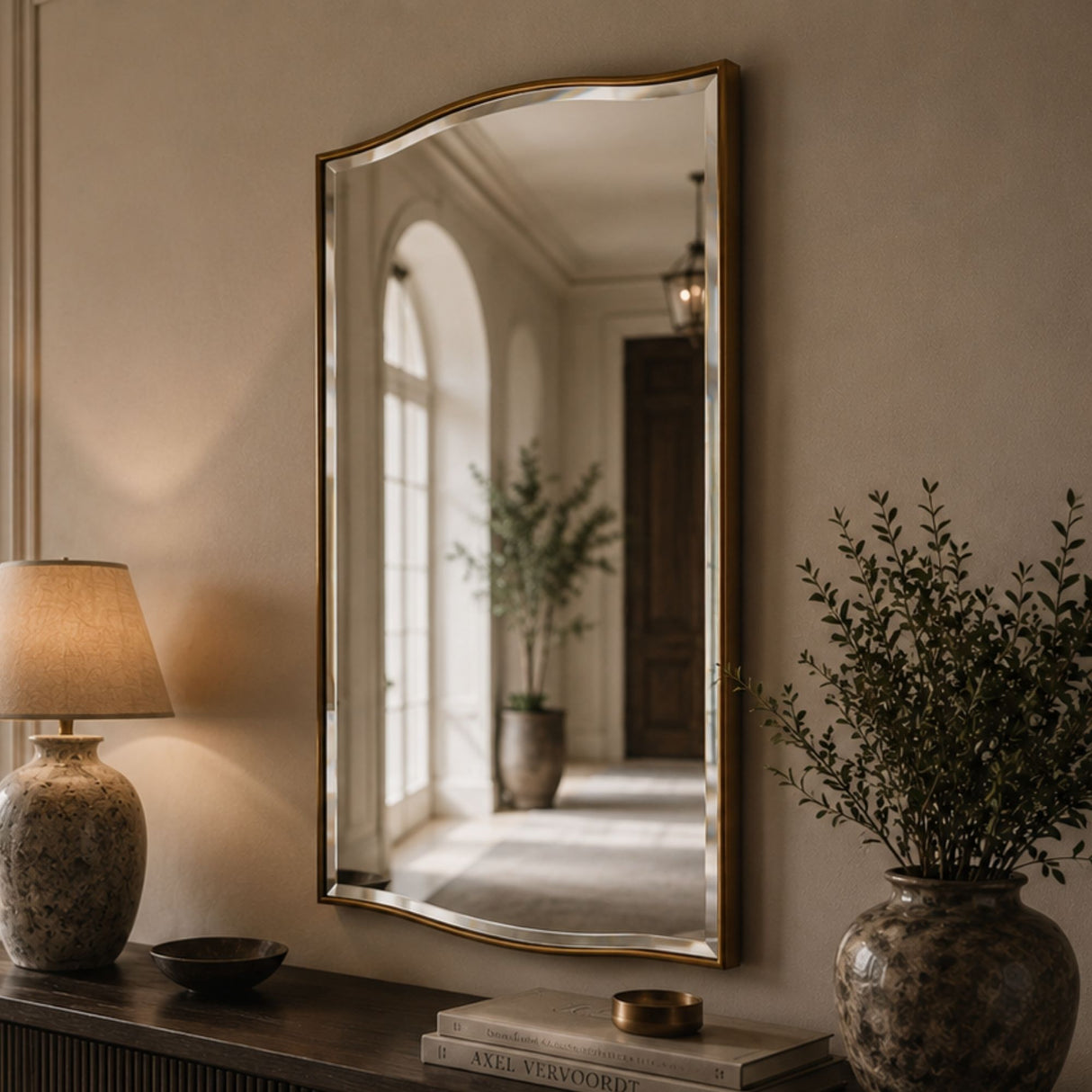 A large arched wall mirror with a gold frame hangs on a beige wall above a console table. The mirror reflects a softly lit hallway with an arched window and potted plants.