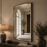 A large arched wall mirror with a gold frame hangs on a beige wall above a console table. The mirror reflects a softly lit hallway with an arched window and potted plants.