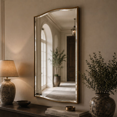 A large arched wall mirror with a gold frame hangs on a beige wall above a console table. The mirror reflects a softly lit hallway with an arched window and potted plants.