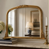 An ornate gold-framed wall mirror with a decorative carved top and corner accents, displayed on a marble console table in a styled interior setting.