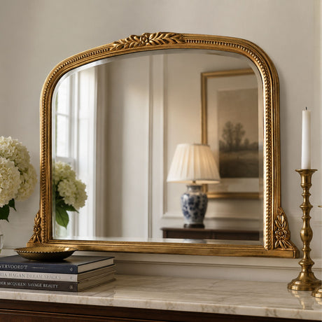 An ornate gold-framed wall mirror with a decorative carved top and corner accents, displayed on a marble console table in a styled interior setting.