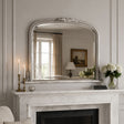 An ornate silver-framed mirror with a decorative leaf crest at the top, mounted on a white marble fireplace mantel in a classic room setting.