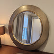 Round gold-framed mirror on a wall with a lamp and window reflection.