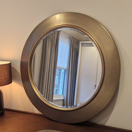 Round gold-framed mirror on a wall with a lamp and window reflection.