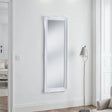 White-framed mirror on a white wall with a floor lamp in a minimalistic room.