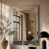 A large rectangular wall mirror with a polished silver frame hangs on a beige wall in a styled interior. The mirror reflects a window, curtains, and a side table, while the room itself features a vase with branches, a black dome lamp, and a dark bowl on a console table.