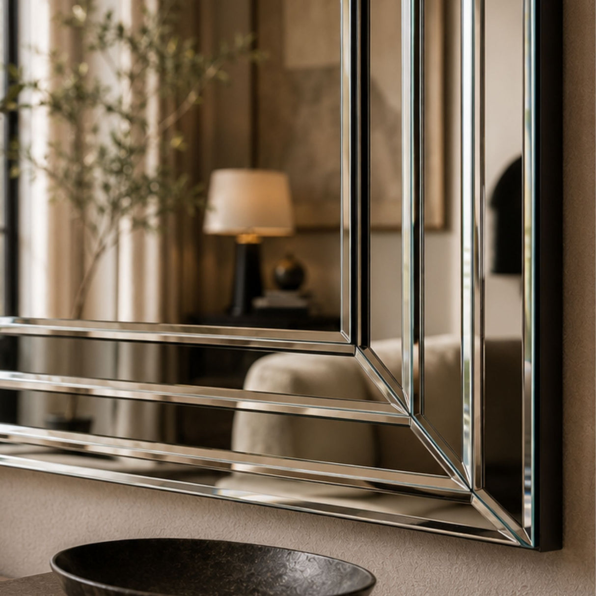 A large rectangular wall mirror with a polished silver geometric frame, mounted on a beige wall above a dark stone vessel sink. The mirror reflects a softly lit modern interior with a lamp and curtains.