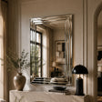 A large rectangular wall mirror with a geometric, beveled silver frame hangs on a beige paneled wall above a console table. The mirror reflects a window and the room interior, while the surrounding scene includes a ceramic vase with dried branches, books, and a black dome table lamp.