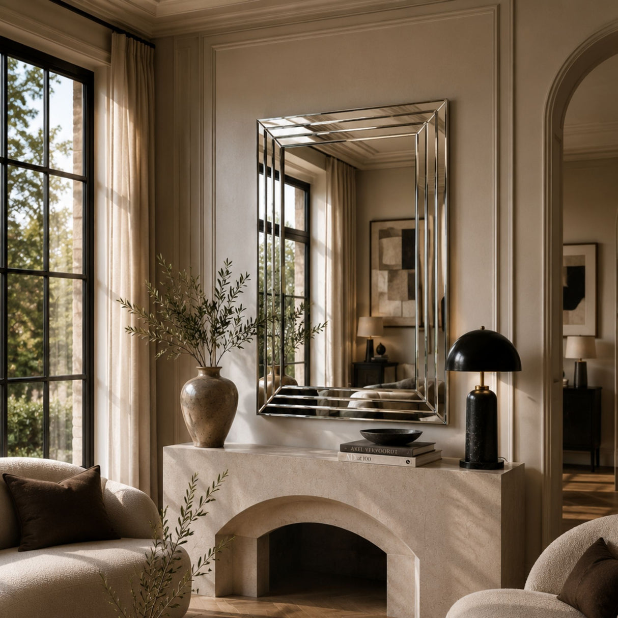 A large rectangular silver mirror with a beveled edge is mounted on a beige wall above a stone fireplace mantel in a styled living room. The room features warm neutral tones, beige curtains, a black table lamp, and a ceramic vase with dried branches.