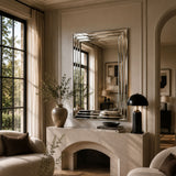 A large rectangular silver mirror with a beveled edge is mounted on a beige wall above a stone fireplace mantel in a styled living room. The room features warm neutral tones, beige curtains, a black table lamp, and a ceramic vase with dried branches.