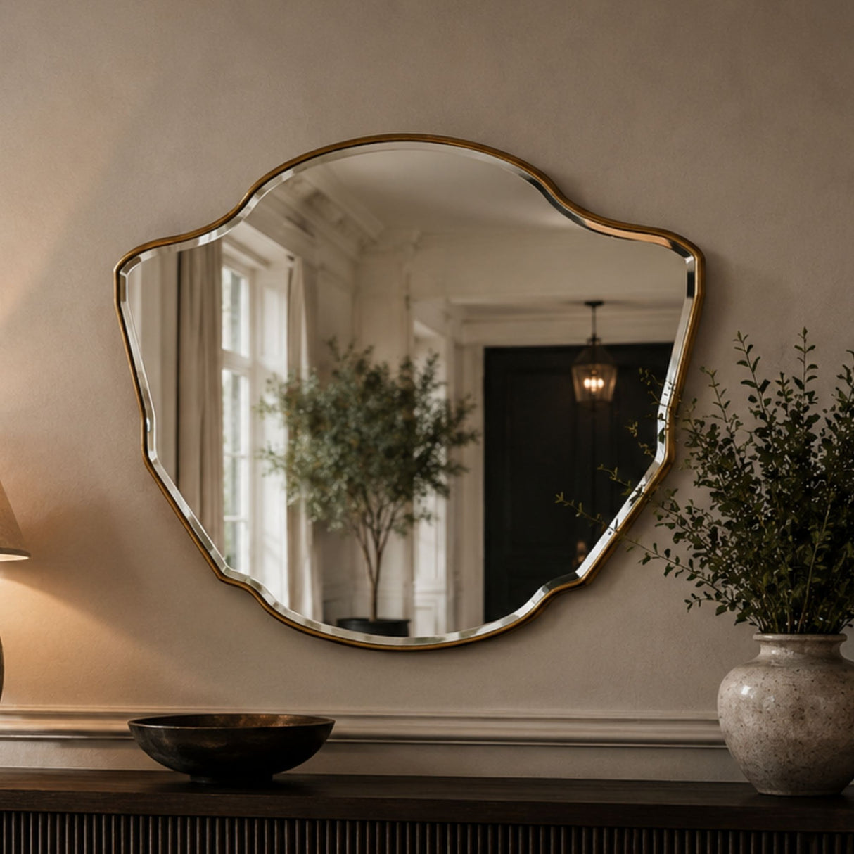 An organic-shaped wall mirror with a gold-toned scalloped frame, mounted on a beige wall above a console table. The mirror reflects a home interior with a tree and pendant light, and the scene is styled with a lamp, bowl, and potted plant.