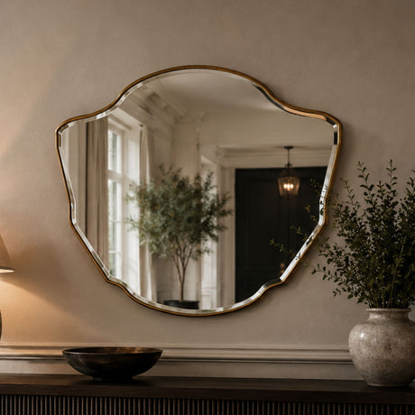 An organic-shaped wall mirror with a gold-toned scalloped frame, mounted on a beige wall above a console table. The mirror reflects a home interior with a tree and pendant light, and the scene is styled with a lamp, bowl, and potted plant.