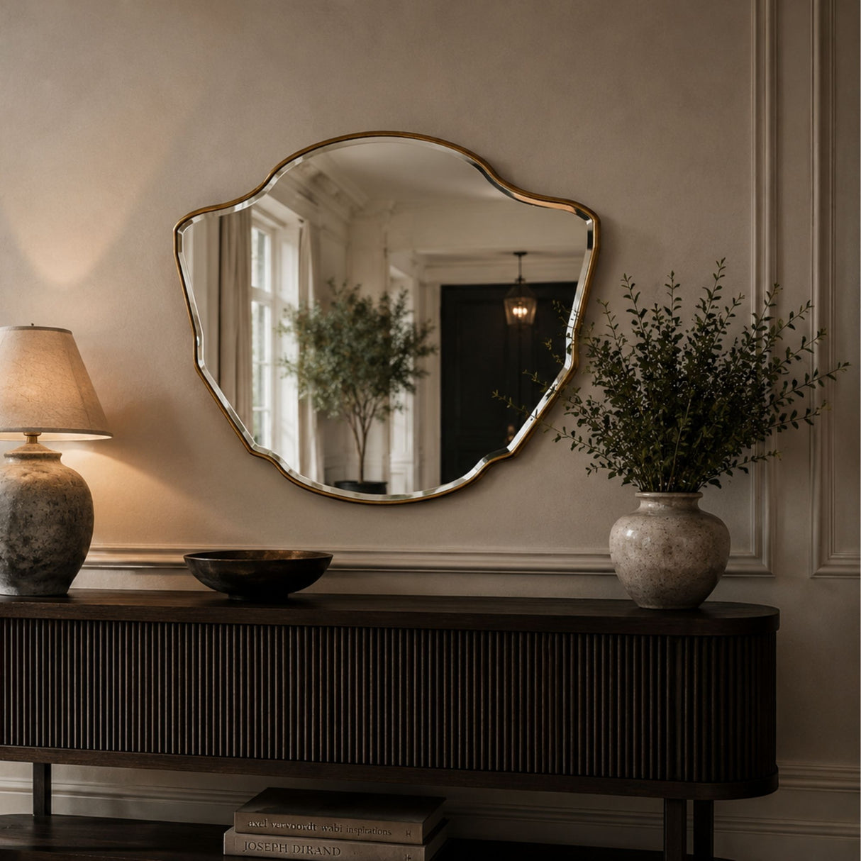 An organic-shaped wall mirror with a gold-toned frame hangs above a dark wood console table in a styled interior. The mirror reflects a room interior, while the table is decorated with a lamp, a bowl, and a vase of greenery.