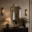 An ornate scalloped wall mirror with a gold-toned frame hangs on a beige wall above a dark wood console table. The room is styled with a lit table lamp, a vase of greenery, and matching wall sconces.