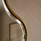 Close-up of a modern wall mirror with a flowing, wavy organic shape and a brushed gold metal frame against a warm beige wall.