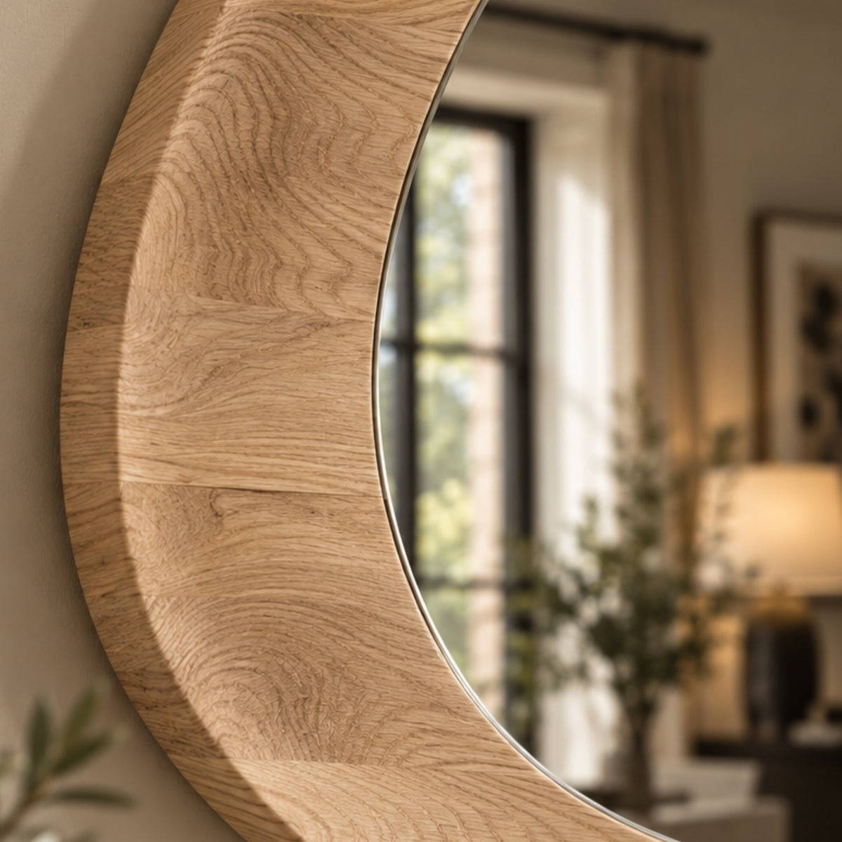 A close-up of a round wall mirror with a thick, curved natural oak wood frame, reflecting a softly blurred interior room with a window, curtains, and a lamp.