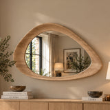 An organic-shaped wall mirror with a thick, natural oak wood frame, mounted on a beige wall above a wooden console table. The mirror reflects a softly lit room interior, and the scene is styled with books, bowls, and a potted plant.