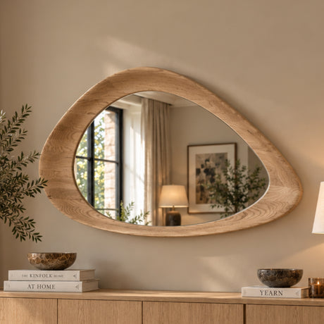 An organic-shaped wall mirror with a thick, natural oak wood frame, mounted on a beige wall above a wooden console table. The mirror reflects a softly lit room interior, and the scene is styled with books, bowls, and a potted plant.